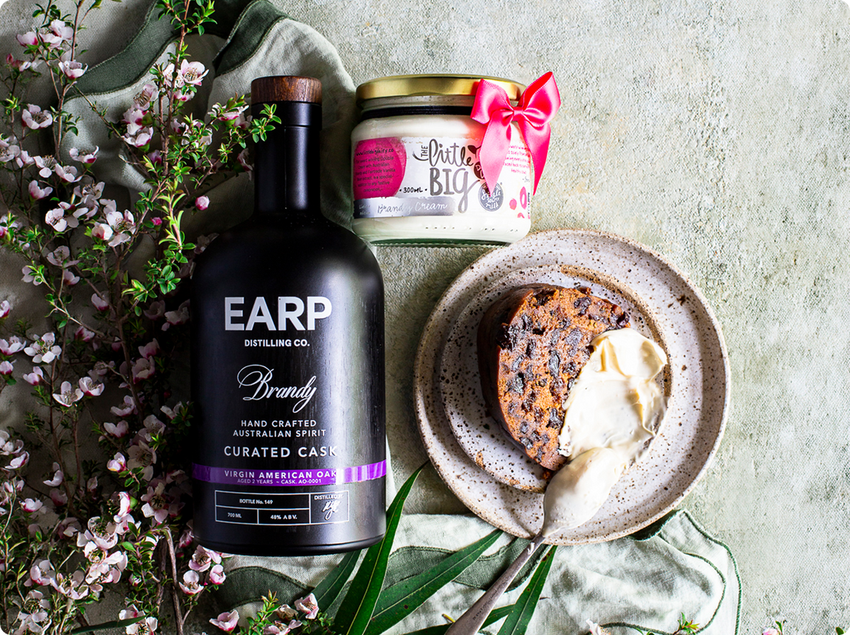 A styled flat-lay featuring a bottle of EARP Distilling Co. and a jar of 'brandy cream', arranged with a slice of fruit cake on a speckled plate with vanilla ice cream, alongside green leaves and pink blossoms on a textured light grey background.