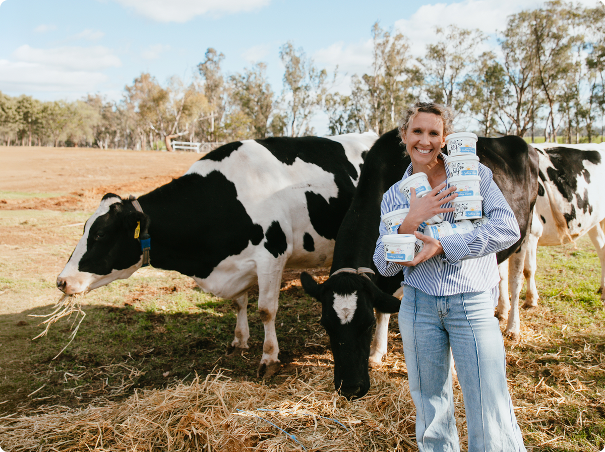 A woman standing on a farm holding several cartons of Little Big Dairy Cottage Cheese with black-and-white cows in a paddock behind her under a blue sky.
