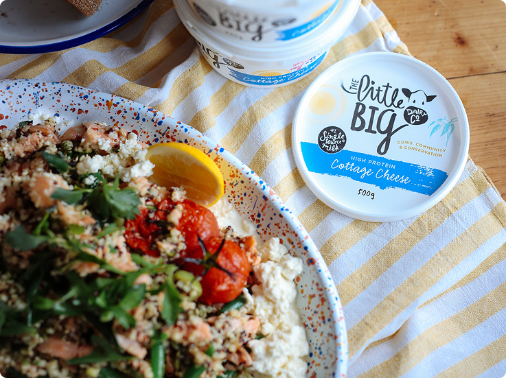 A close-up of a plated meal with couscous, feta cheese, greens, tomatoes, and a lemon wedge beside a container of Little Big Dairy Cottage Cheese. The product lid is placed on a striped cloth over a wooden table.