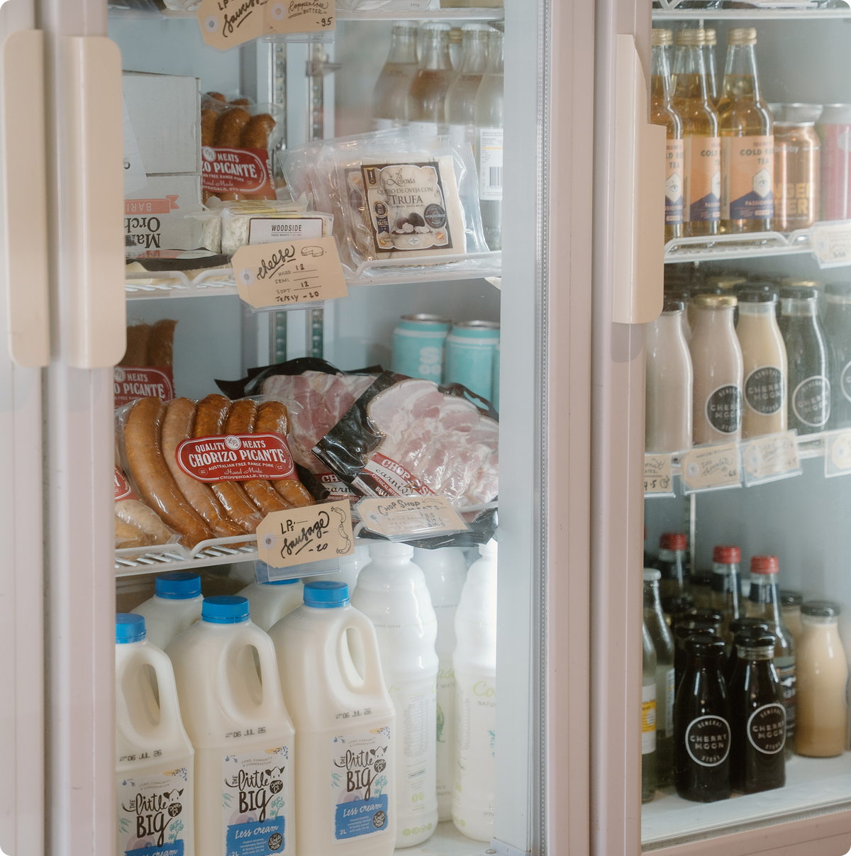 A well-stocked refrigerator with two glass doors, filled with milk cartons, packaged meats, bottled beverages, and other grocery items. Hand-written price tags are attached to several items.