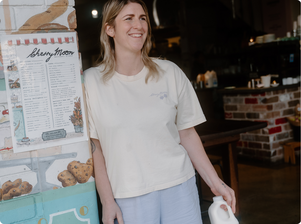 A woman smiling in a cafe setting, wearing a cream-coloured t-shirt with a small light blue graphic on the left chest and light blue pants, holding a white plastic milk bottle.