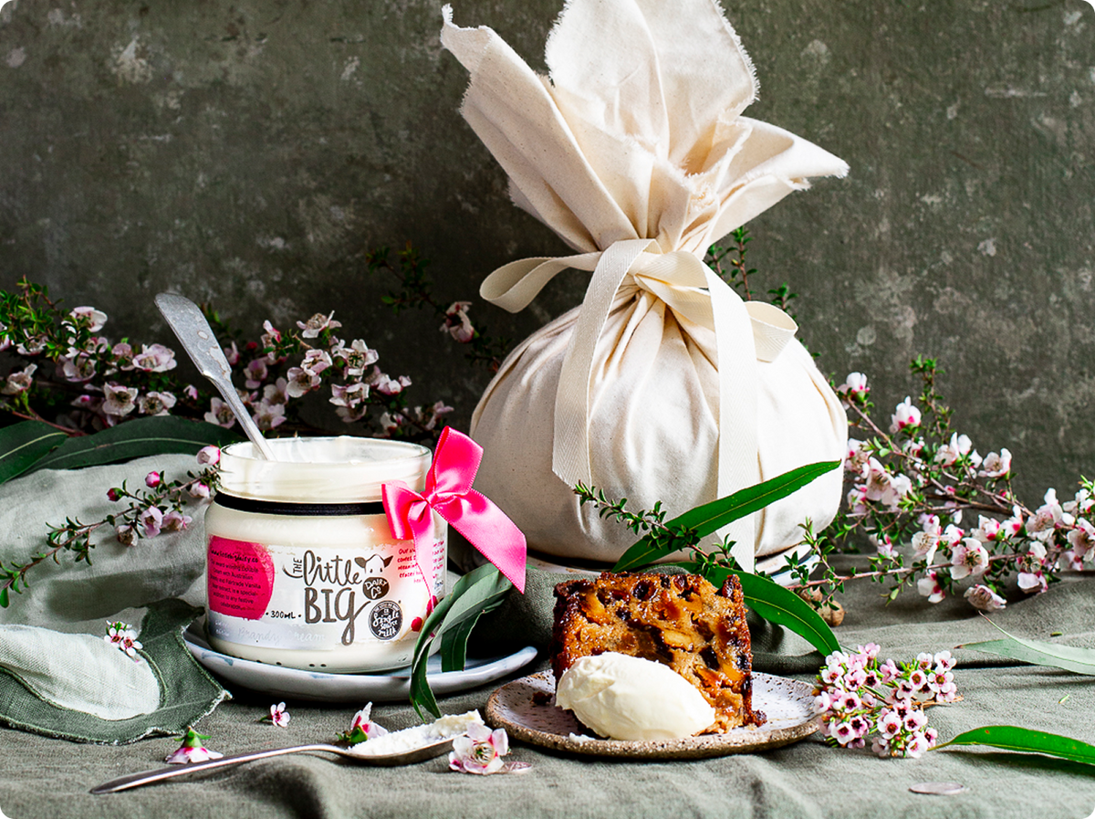 A styled product presentation featuring a jar of brandy cream, a large cream fabric gift bag tied with a satin ribbon, and a slice of fruit cake, all arranged with green leaves and small pink flowers against a textured grey background.