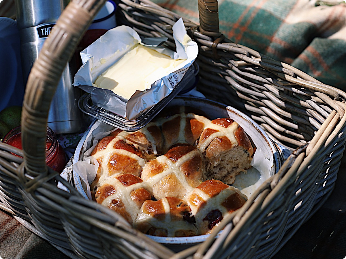 A wicker picnic basket filled with baked hot cross buns, a tin of butter on foil, and other picnic items, set outdoors on a plaid blanket in bright sunlight.