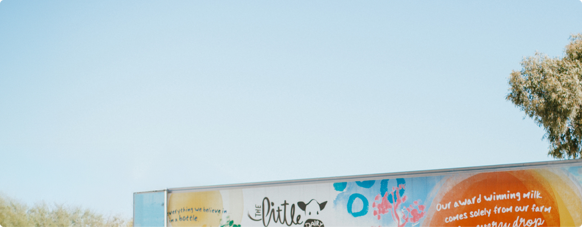 A close-up of a Little Big Dairy branded truck against a clear blue sky, with part of a tree visible on the right. The sign includes stylised text and abstract blue, pink, and yellow graphics.