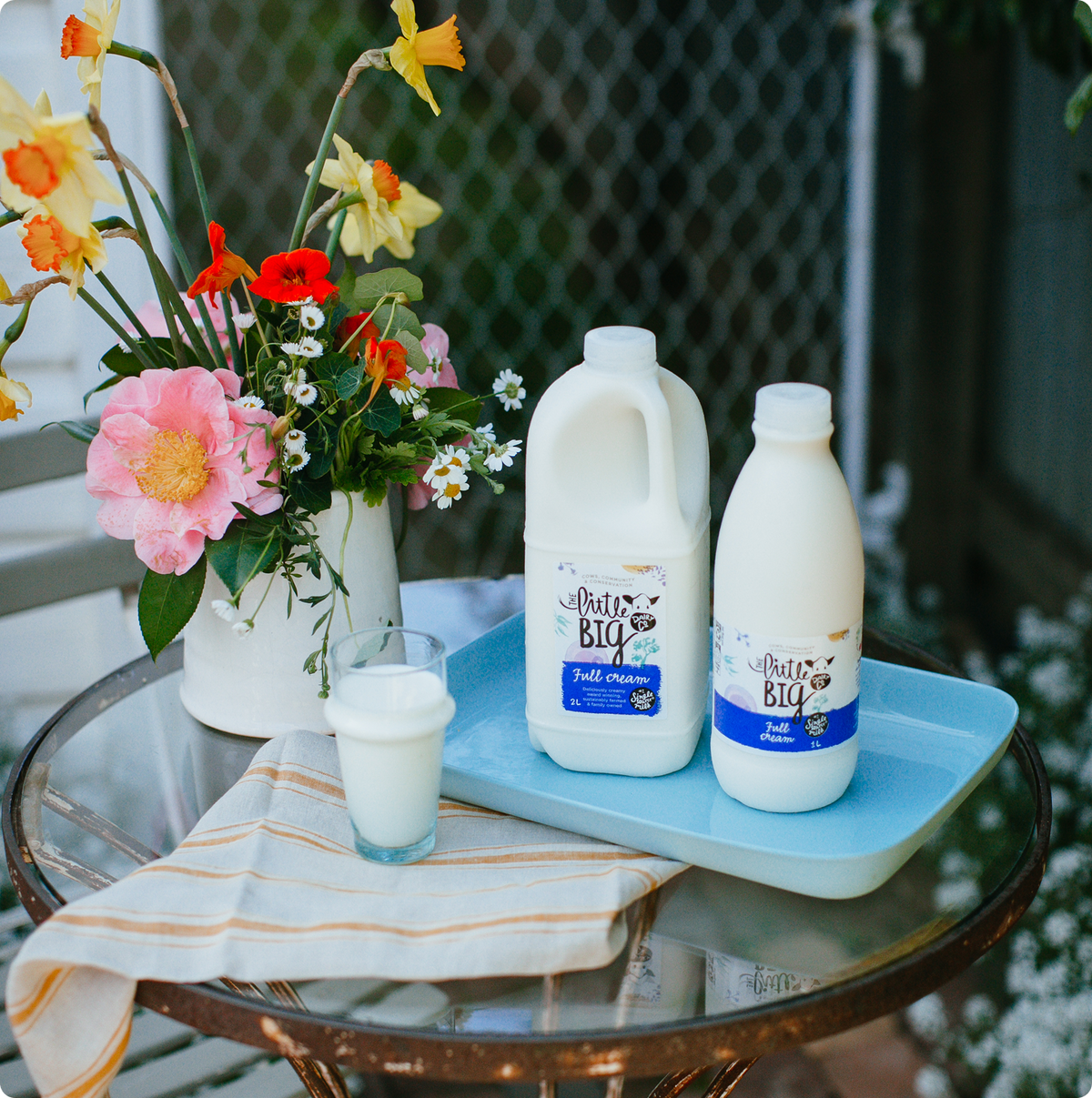 Two milk products from Little Big Dairy on a light blue tray, displayed on a glass table next to a glass of milk, a vase of colourful flowers, and a striped napkin in an outdoor patio setting.