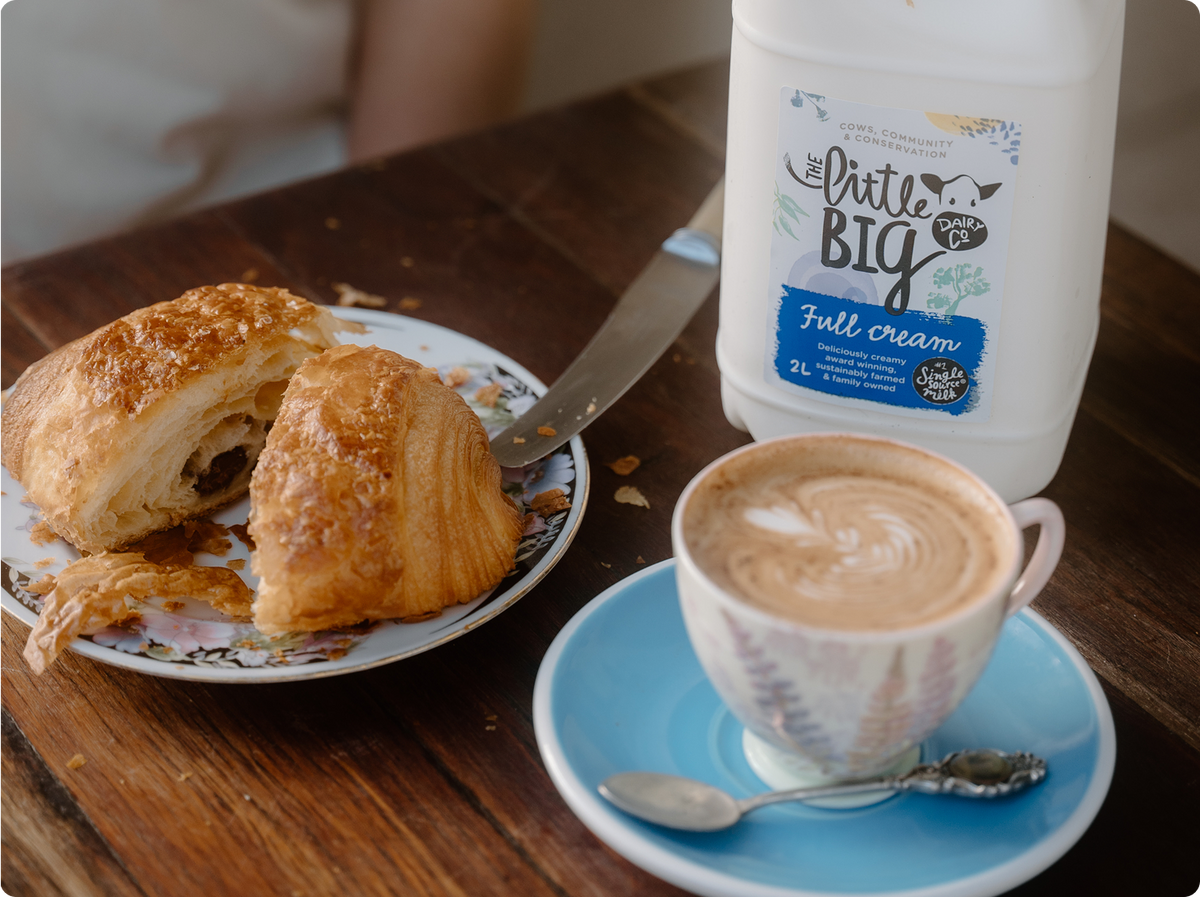 A cup of latte art coffee on a blue saucer next to a pastry on a plate and a bottle of Little Big Dairy Full Cream milk on a dark wooden table.